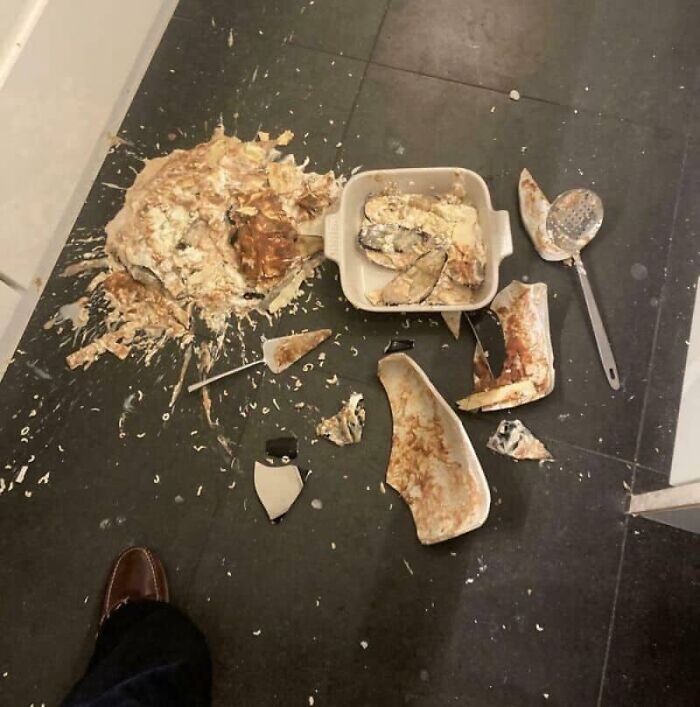A catastrophic kitchen fail with a broken casserole dish and food splattered on a dark tile floor.