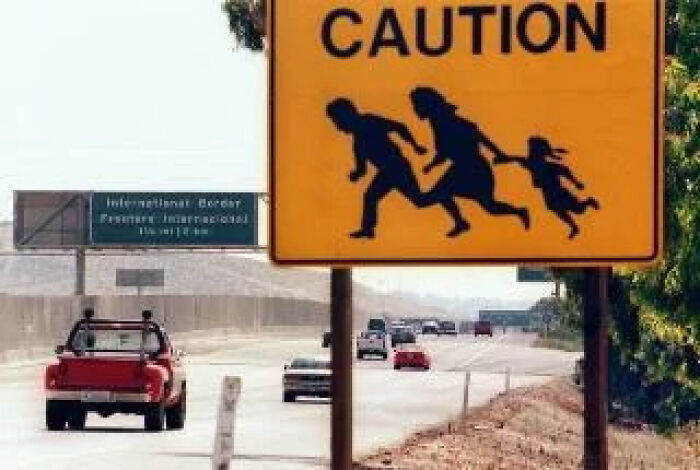 A yellow Caution road sign depicting a running family next to a highway with passing cars