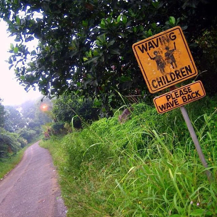 A yellow roadside sign saying Waving Children Please Wave Back, showing funny mistakes on signs