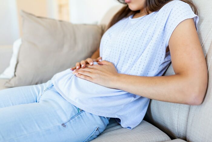 A woman sitting on a couch holding her stomach, dealing with sudden body changes in her 30s and 40s.