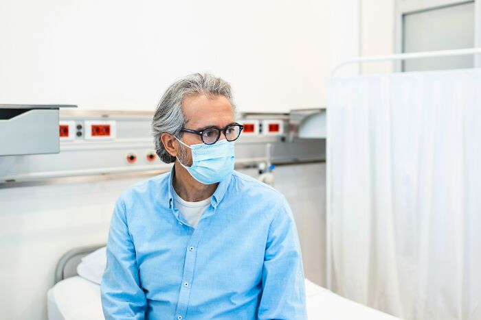 A man wearing a mask sits on a hospital bed, looking concerned about standard medical procedures.