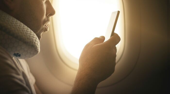 A passenger wearing a neck pillow and using his phone while sitting next to a plane seatmate on a flight.