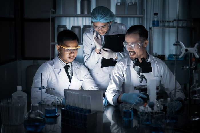 Three scientists in a dimly lit lab examining a laptop and microscope, uncovering industry secrets through research and development.