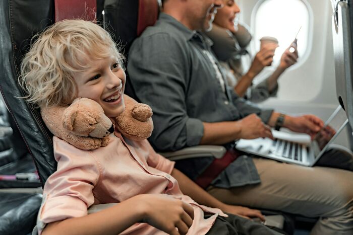 A smiling boy with a teddy bear neck pillow sits next to his father on a plane, avoiding a plane seatmate from hell.