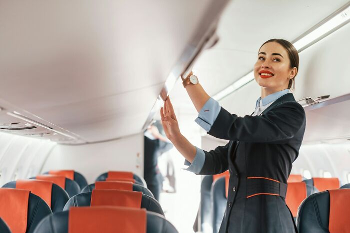 A smiling flight attendant standing by the cabin seats to help a plane seatmate with overhead luggage.