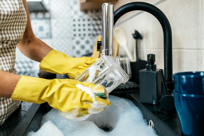 Hands wearing yellow rubber gloves washing a blender cup in a sink, showing cleanup after events like weddings.