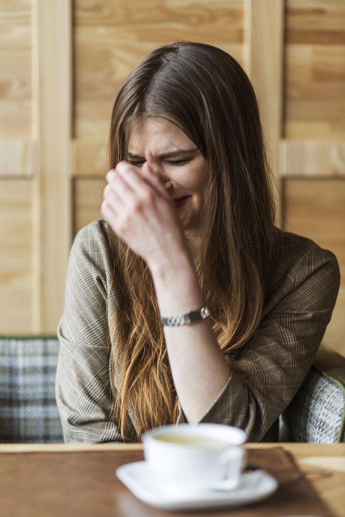 A young woman laughing and covering her face at a table with a cup of tea, possibly after overhearing something.