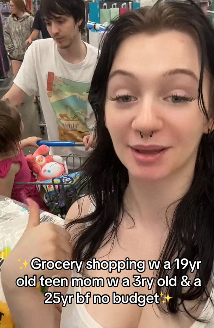 American parents grocery shopping with a full cart, featuring a woman with a child and man behind her. American parents grocery shopping with a full cart, featuring a woman with a child and man behind her.