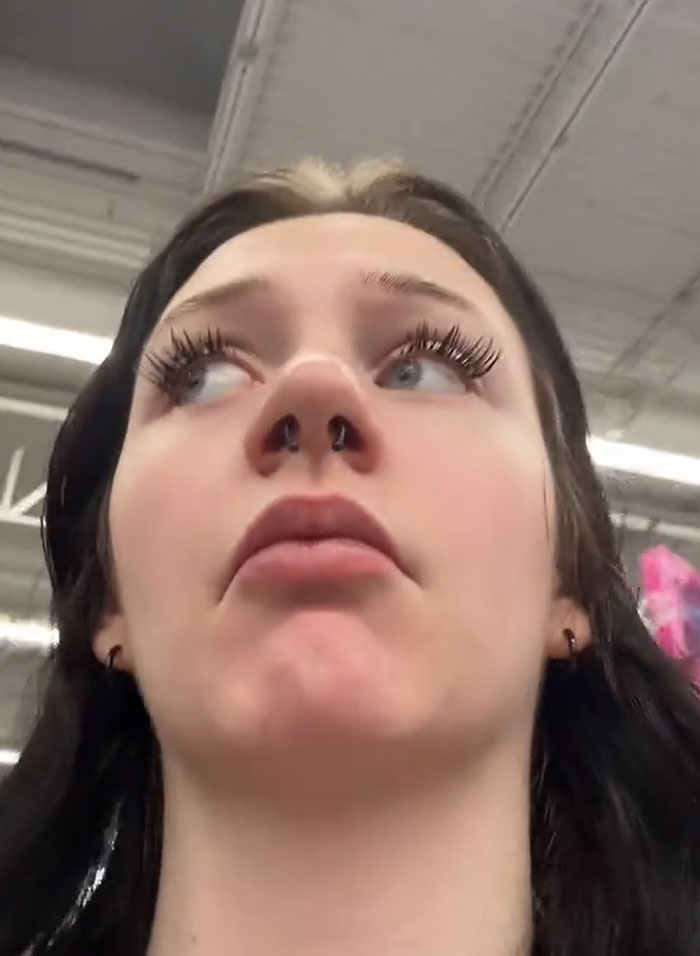 A young woman with long eyelashes and a septum piercing looks up, expressing concern during groceries shopping. A young woman with long eyelashes and a septum piercing looks up, expressing concern during groceries shopping.