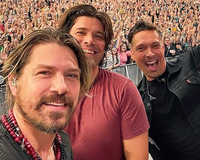 Three Hanson brothers smiling in front of a large excited crowd at a live concert