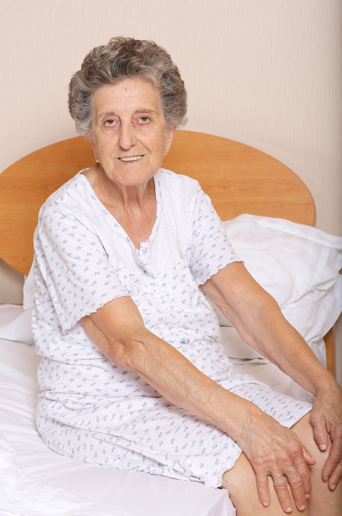 An elderly woman in nightwear sitting on a bed, potentially showing pain reactions.