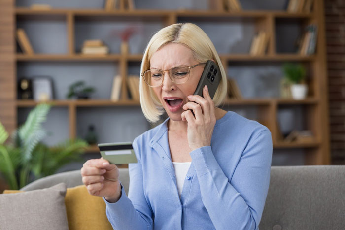 A wife looks shocked while on the phone, holding a credit card due to her husband's spending and using her information.