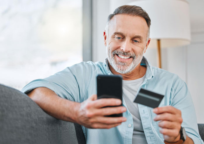 A smiling man on a couch, holding a phone and a credit card, related to a wife separating finances due to husband's spending.