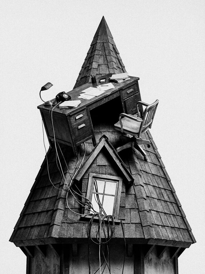 An office desk and chair precariously balanced on a tall, conical roof, creating a surreal and dystopian image.
