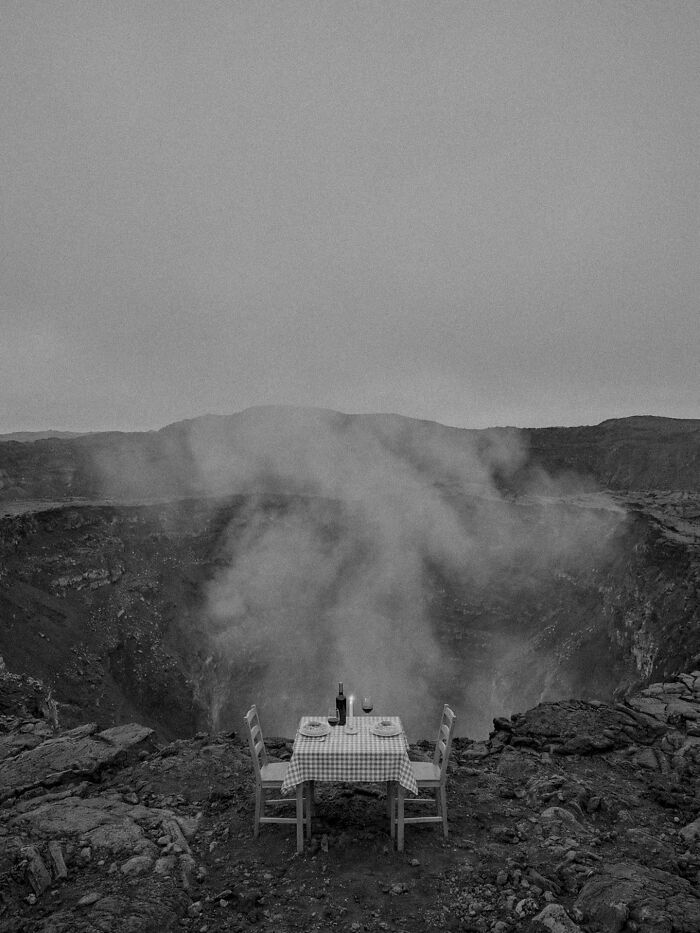 A small table set for dinner at the edge of a smoking volcanic crater, a surreal and dystopian image.