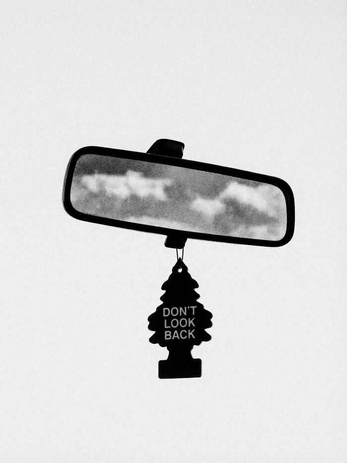 A car rearview mirror reflecting clouds, with a tree-shaped air freshener stating 'Don't look back', a surreal image.