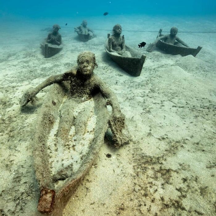 Underwater sculptures of figures in boats on the ocean floor, forming an art gallery for marine life.