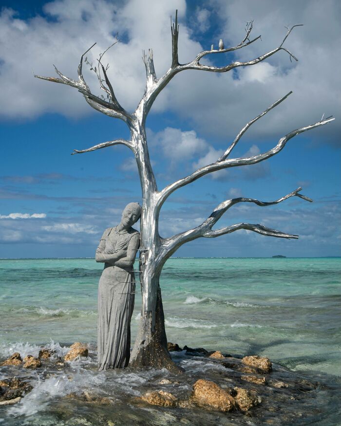 A woman sculpture leaning against a tree on an ocean floor, showcasing underwater sculptures and supporting marine life.