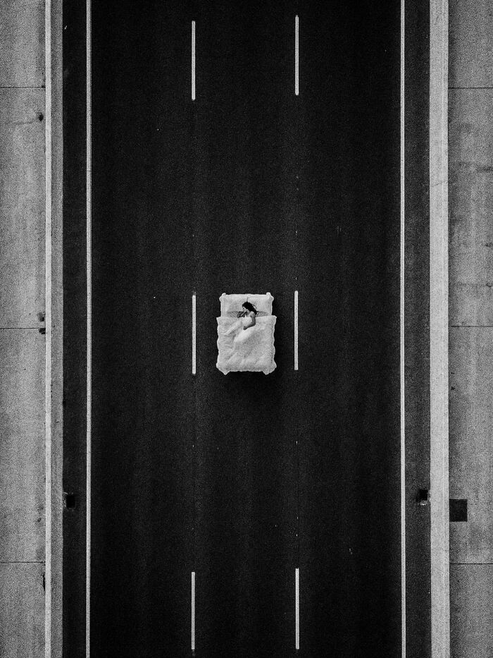 A black and white aerial view of a person sleeping in a bed in the middle of a desolate road, a surreal image by Marcus Wallinder.
