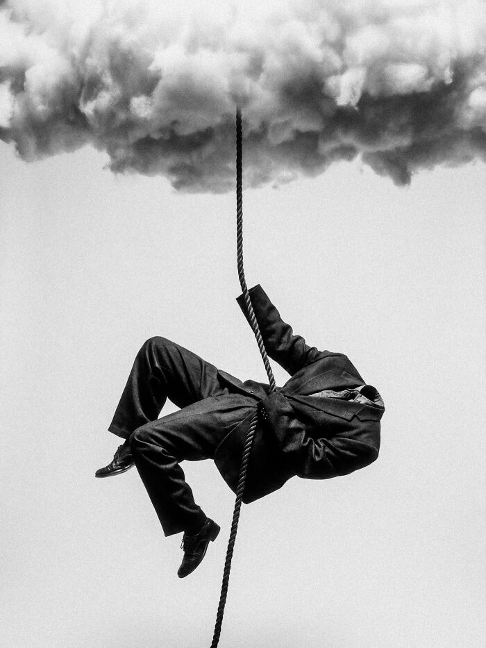 A black and white image of a headless man in a suit climbing a rope leading into a surreal cloud, symbolizing distorted reality.