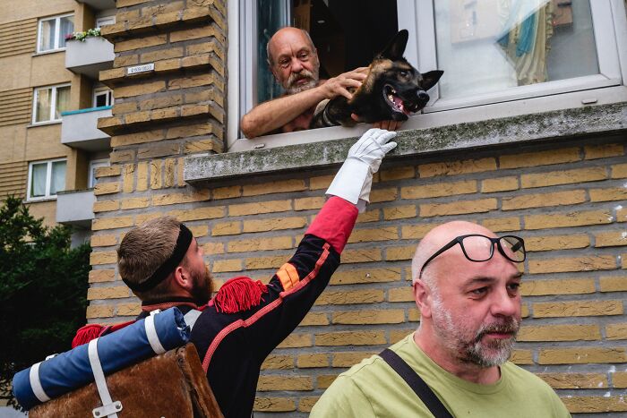Jeffrey De Keyser's street photography depicts a man holding a dog from a window, interacting with people below.