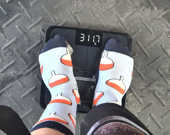 A person in fishing bobber socks stands on a digital scale displaying 311.7 lbs, highlighting a weight loss transformation. A person in fishing bobber socks stands on a digital scale displaying 311.7 lbs, highlighting a weight loss transformation.