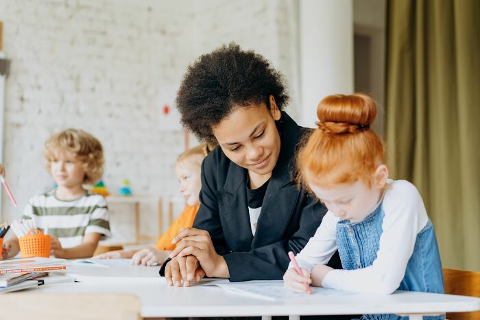 A teacher helps a young girl with red hair write at a desk, surrounded by other children, representing industries that are about to collapse.