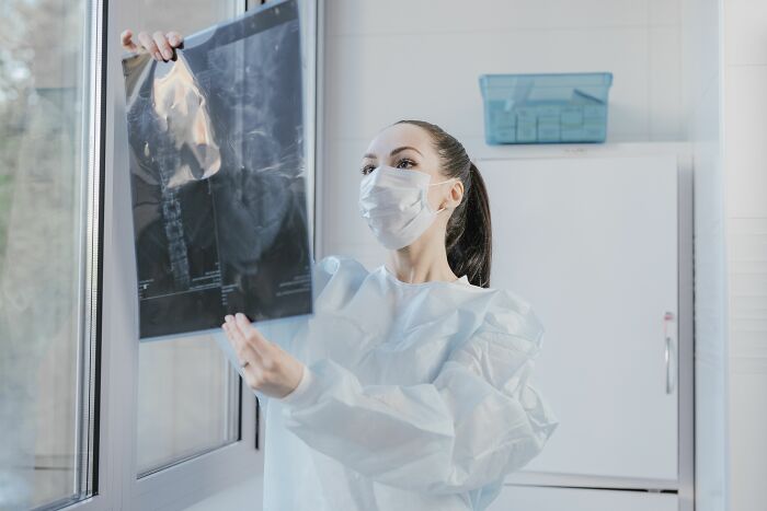A female doctor in a mask and gown holds up an X-ray image, possibly from a collapsing industry.