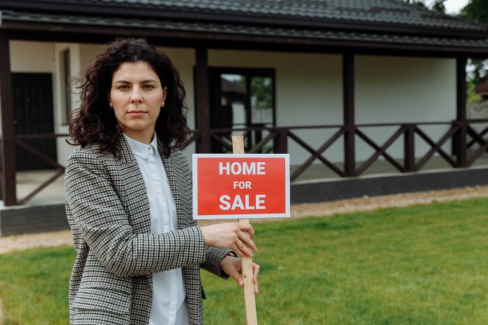 A woman holding a Home for Sale sign, representing industries that are about to collapse, like real estate.
