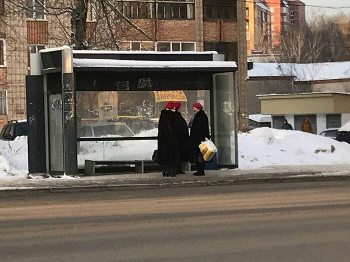 Two women in red hats and black coats standing at a snowy bus stop, synced just right with their surroundings.