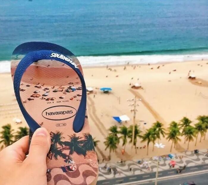 A hand holds a flip-flop featuring a beach scene, perfectly synced with the real beach view below.