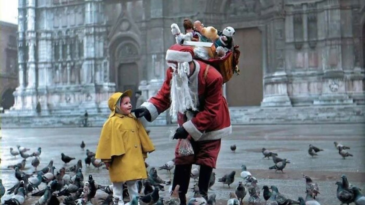 Historical colorized photo: Santa Claus with a bag of toys interacts with a small child in a yellow coat, surrounded by pigeons.