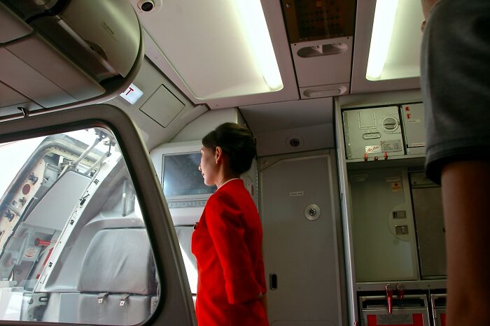 A cabin crew member in a red uniform stands in the galley of an airplane, ready to deal with passenger requests.