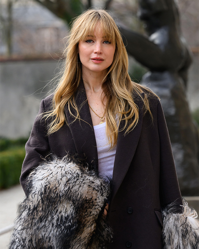 Jennifer Lawrence with long blonde hair and bangs, wearing a dark coat and holding a furry garment. Her plastic surgery confession sparks scrutiny.