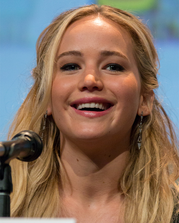 Jennifer Lawrence speaking at a microphone, smiling broadly with long blonde hair. Her plastic surgery confession sparks scrutiny.