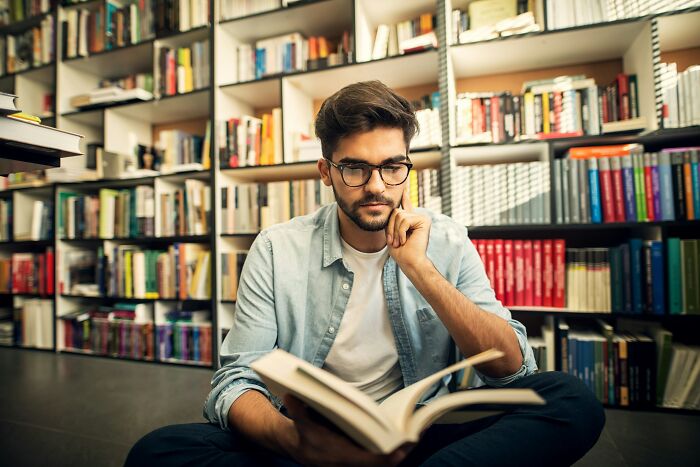 Young man reading a book in a library, reflecting on his career options in a difficult job market.