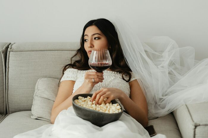 A stressed bride in her wedding dress on a couch eating popcorn and holding wine during wedding planning mishaps