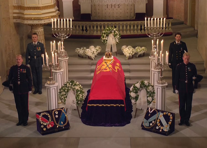 Royal funeral with a crucial role for the Crown Prince, a coffin draped in flag, surrounded by guards and wreaths.