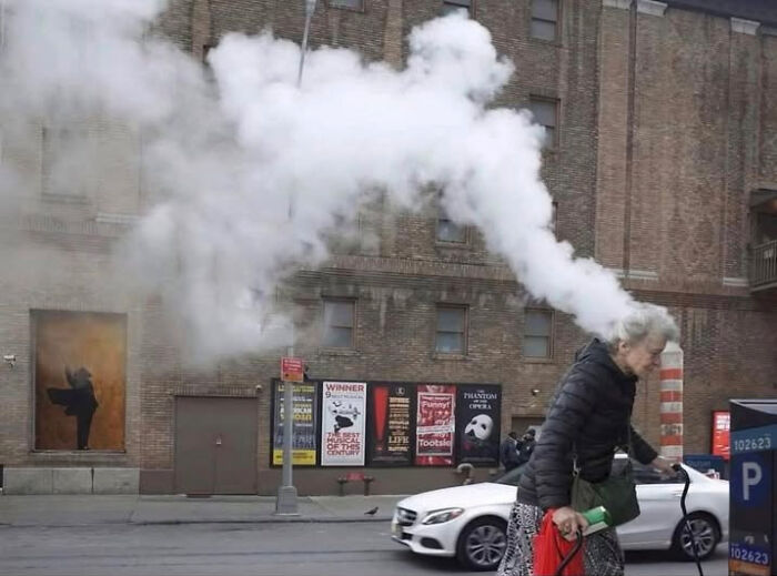 A confusing perspective of steam from a street pipe looking like smoke coming out of an elderly woman's head.