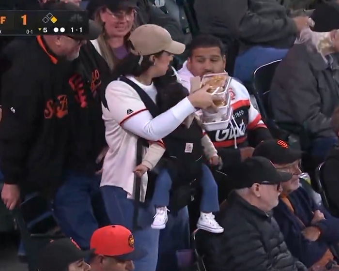 A mother carrying a baby in a carrier and holding food while a man looks on, related to viral baseball match. A mother carrying a baby in a carrier and holding food while a man looks on, related to viral baseball match.