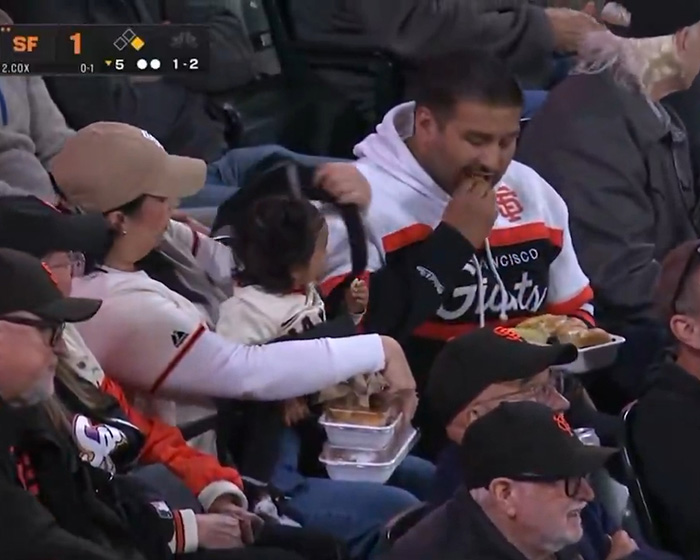 A man eating food from a tray at a baseball game while a woman next to him holds containers, sparking viral debate. A man eating food from a tray at a baseball game while a woman next to him holds containers, sparking viral debate.