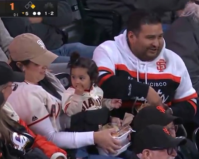 Husband eats while wife holds baby, relating to viral treatment of wife during baseball match. Husband eats while wife holds baby, relating to viral treatment of wife during baseball match.