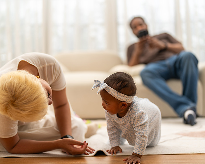 Mother plays with baby on floor while father uses phone, representing viral husband treatment of wife. Mother plays with baby on floor while father uses phone, representing viral husband treatment of wife.