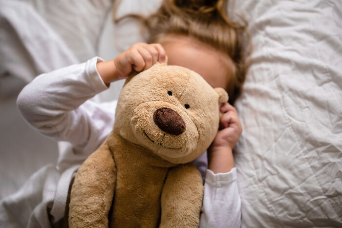 A child lying in bed, holding a teddy bear over their face, symbolizing comforting childhood beliefs.