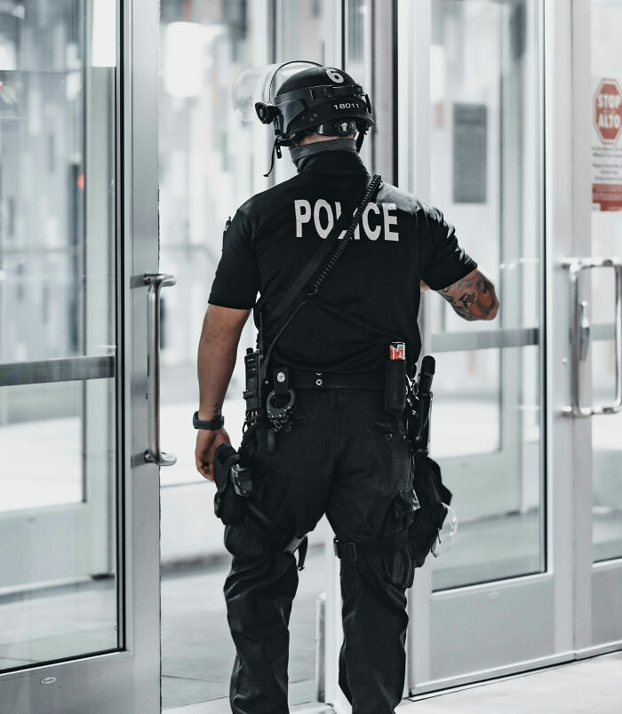 A police officer in riot gear with POLICE on their back stands facing a glass door, unrelated to flight attendants rolling their eyes.
