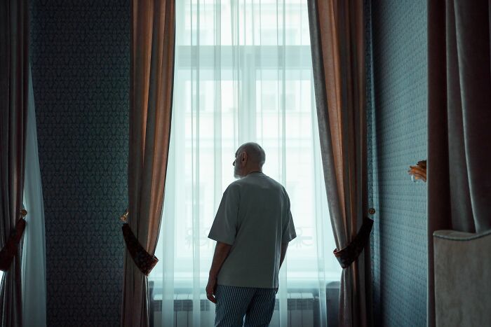 An elderly man stands alone, looking out a window, reflecting on the profound switches in his long marriage.