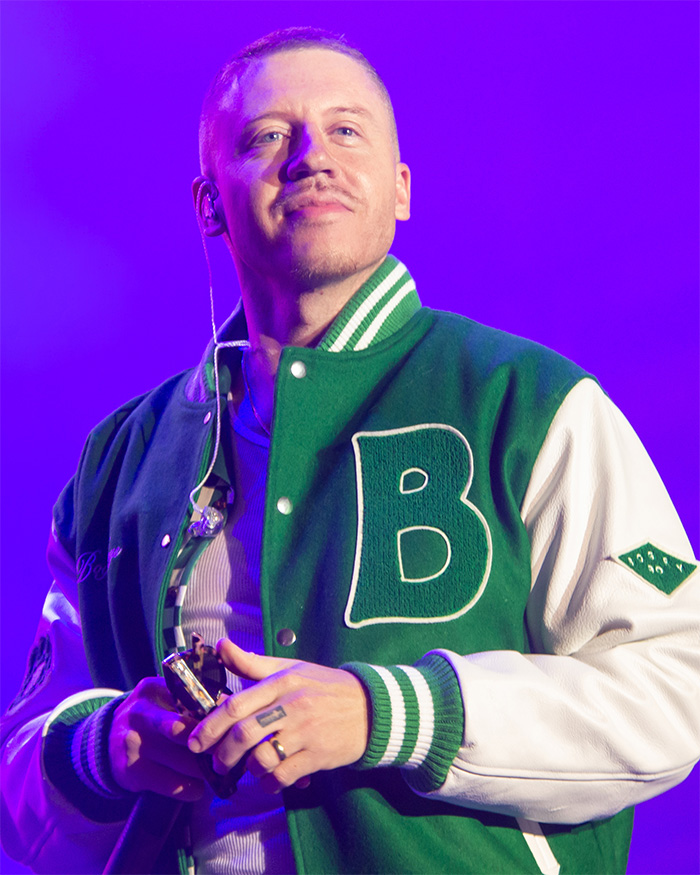 Macklemore performs live in a green and white varsity jacket, looking towards the audience on the Ed Sheeran Tour.
