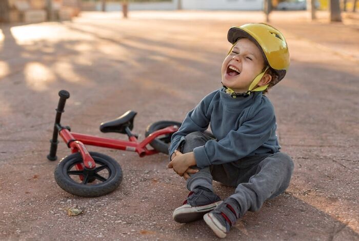 A child with a helmet crying on the ground next to a fallen bicycle, expressing pain reactions.