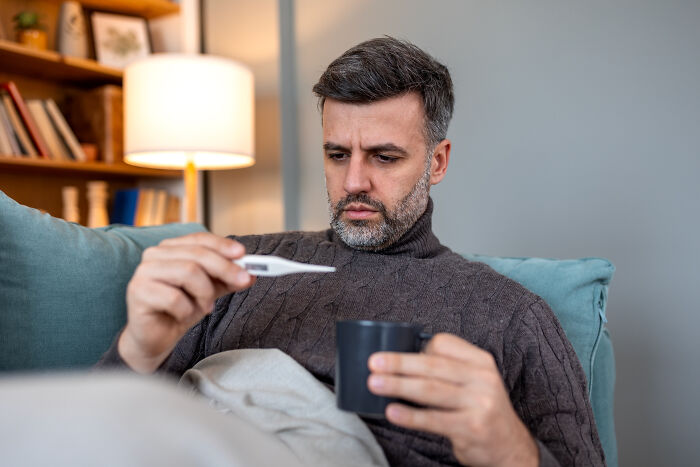 A man with a beard sits on a couch, looking at a thermometer while experiencing illness and sickness.