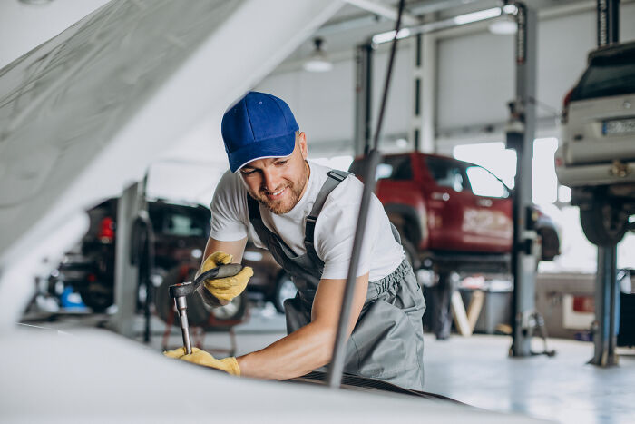 A male auto mechanic smiling while working on a car engine, depicting a job where women face subtle sexism.